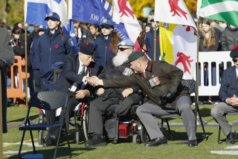 Seated veteran in uniform shake hands in front of flags at a ceremony