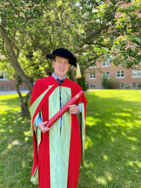 Dr. Kyle Elliott in academic regalia holds his Principal's Prize for Outstanding Emerging Researcher