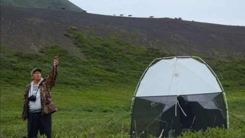 A man raises a weather testing device in the air beside a tent outside, caribou walk along a ridge in the distant background
