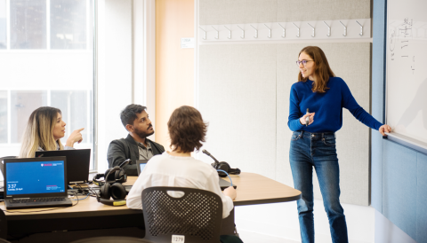 Woman stands in frnt of white speaking to people seated around a boardroom table