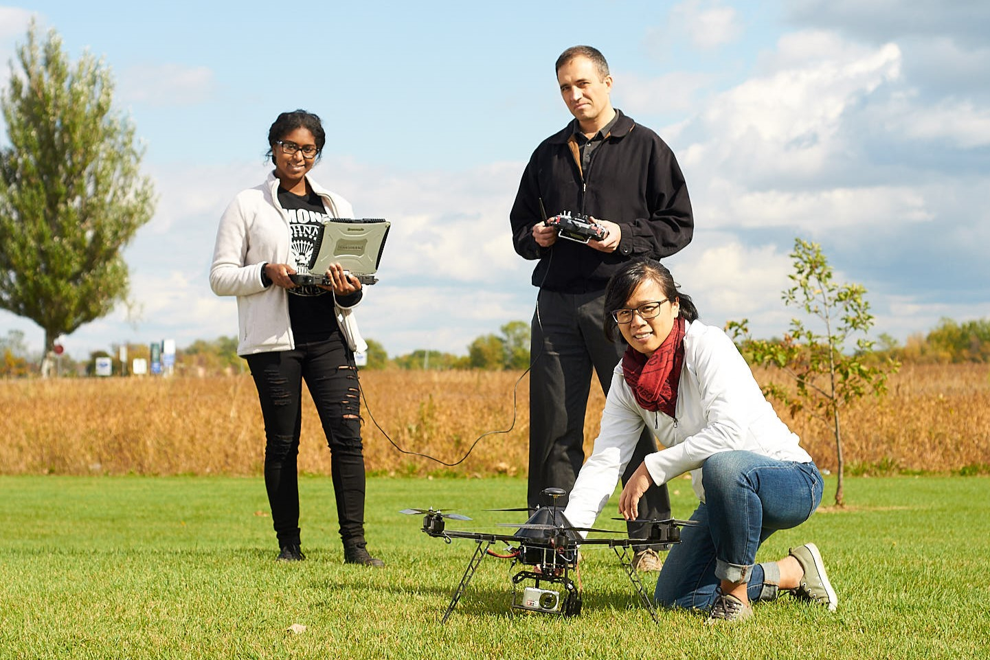 Viacheslav Adamchuk stands in a farm field with two students, one holds a drone, the other holds a laptop