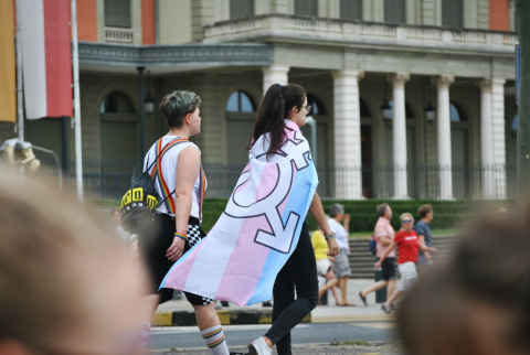 A person walks down a street while draped in a trans flag