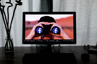 A computer monitor displays a picture of a person looking through binoculars with Facebook logos stuck onto the lenses