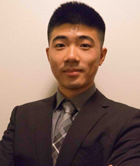Tianqi Wang in suit and tie smiles at camera in front of neutral wall