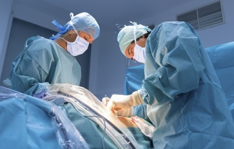 An operating room in which two masked and gowned surgeons perform a procedure on the abdomen of a patient laying on a surgical table