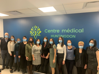 Twelve doctors from the Huntingdon Medical Clinic stand together with masks on, in front of a blue wall with the clinic's logo.