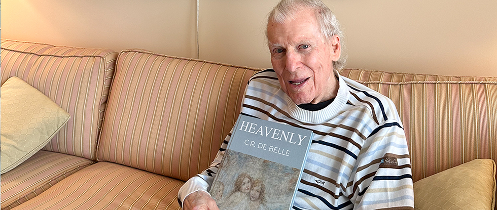 Retired pediatrician Dr. Ross de Belle poses on couch with his book, Heavenly