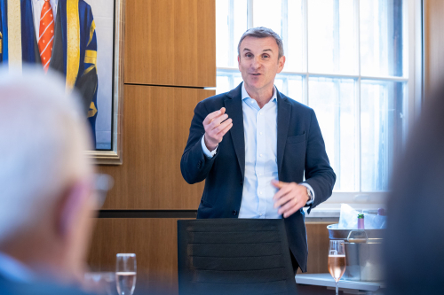 A standing man addresses listeners seated at table in a wood-panelled boardroom.