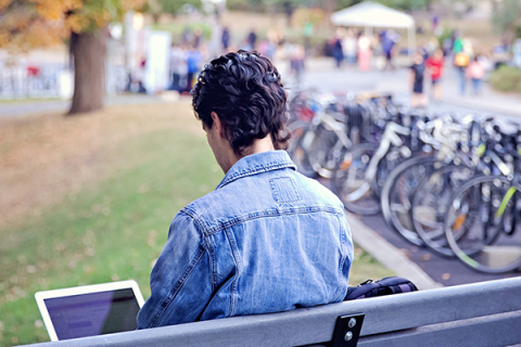 Student sitting on a bench on campus, working on computer