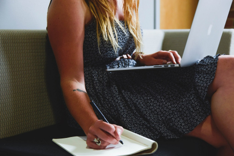 Student writing in a notebook while looking at a laptop