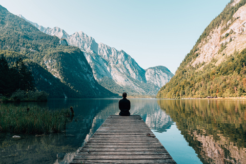 Man sitting at the edge of wood deck, facing lake and surrounding mountains.