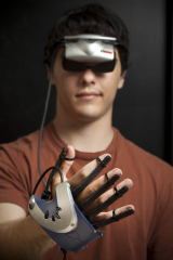 Image of a person wearing a virtual reality headset over their eyes and a glove controller on their hand, holding up their hand in front of their face.