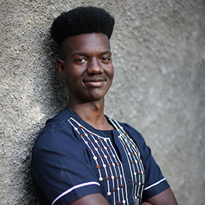Young black man stands against a wall and smils at the camera