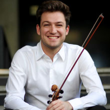 Young man in white shirt sits while smiling at the camera and holding a violin