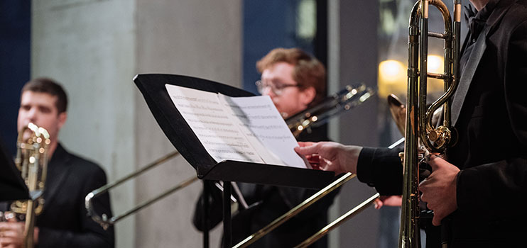 musician holding trombone and reviewing score while other brass musicans play