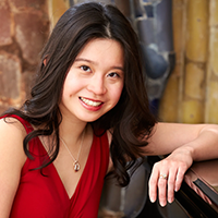 A smiling Nicole Chan wearing a red dress leans one arm on piano