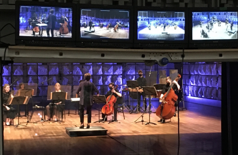 Photo of musicians, including cello, double bass, and percussion being led by a conductor. They are seen through a recording studio window with televisions capturing various angles of the recording session. 