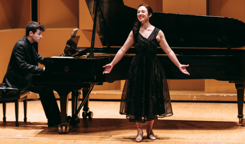 Singer standing in front of piano with arms out, pianist plays piano