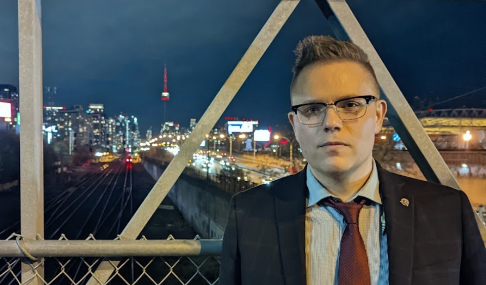 Eliot Britton in a suit and red tie standing on an overpass with the CN tower in the background