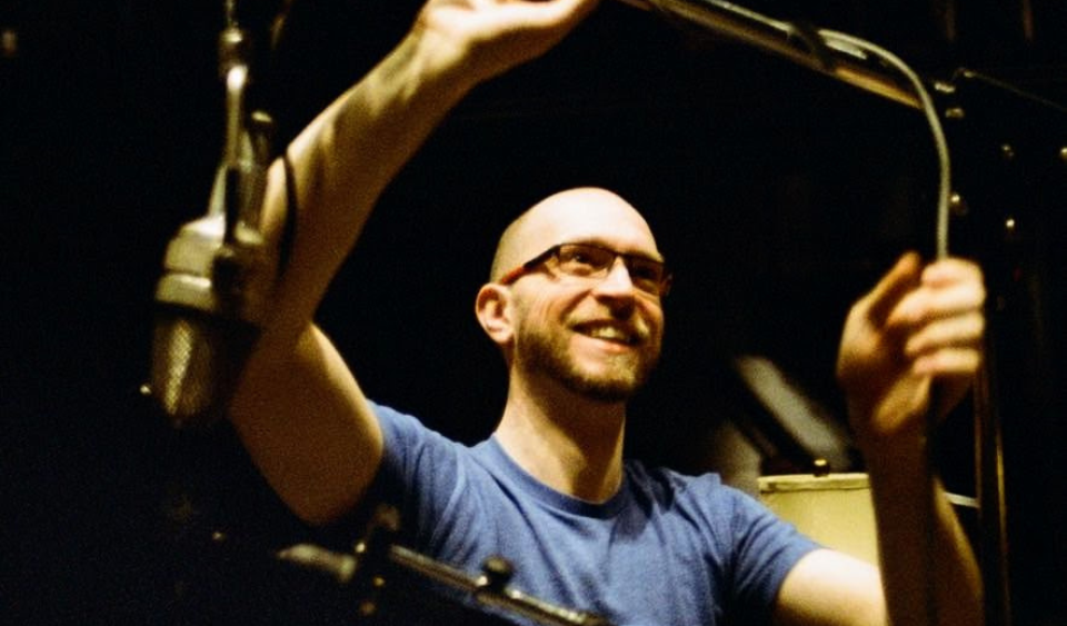 James Clemens-Seely ajuste un microphone dans un studio d’enregistrement, portant des lunettes et un t-shirt bleu.