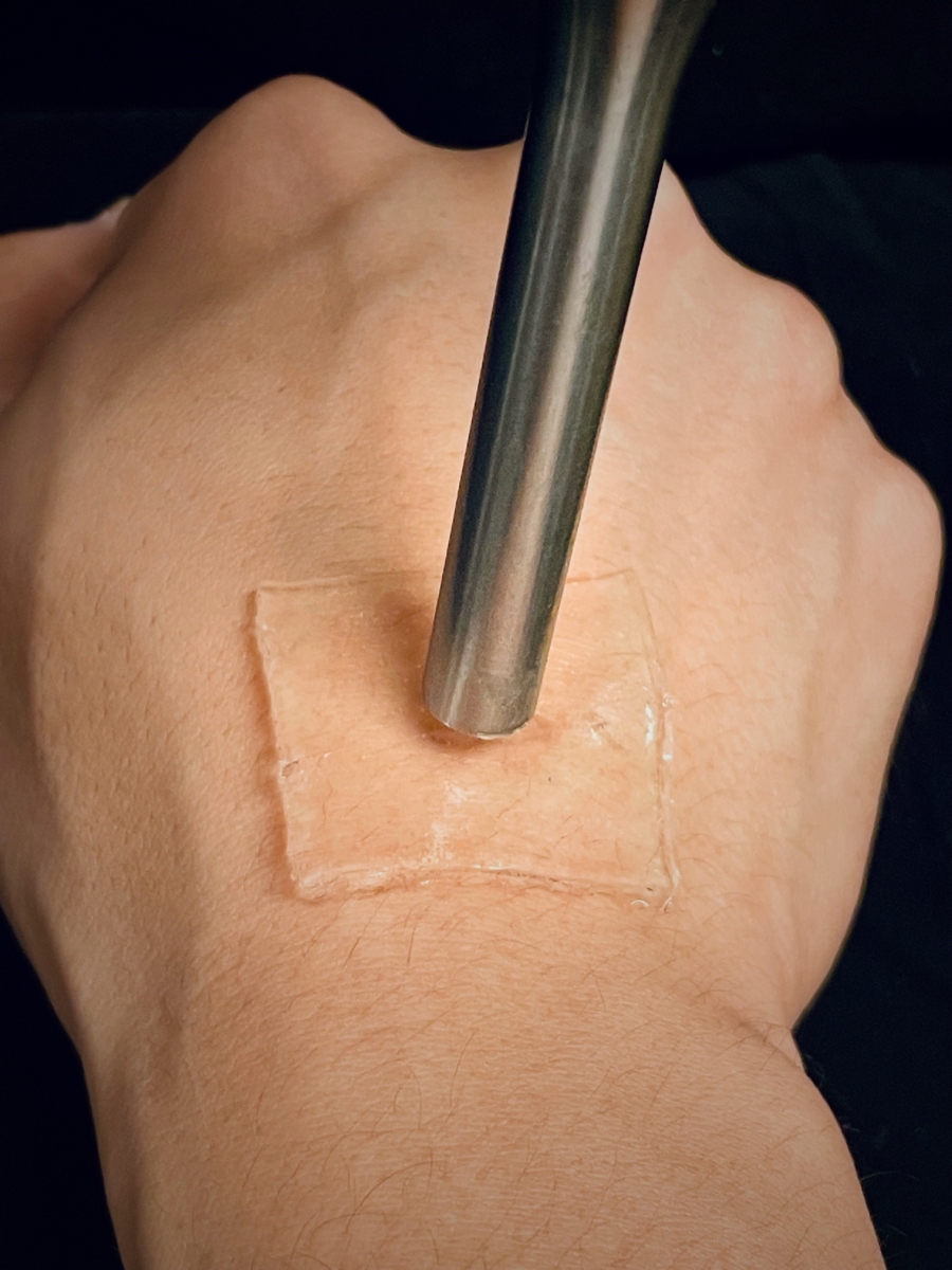 A metal tube hovers over a clear square of adhesive material on the back of a person's hand.