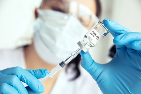 A masked healthcare worker holds a COVID-19 vaccine vial and syringe.