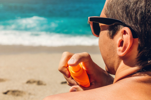 Image: Man on the beach sprays sunscreen on his shoulder. 