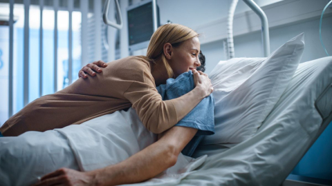 Woman visits a recovering patient in the hospital who is lying in bed. / Femme rendant visite à un patient hospitalisé étendu dans son lit.