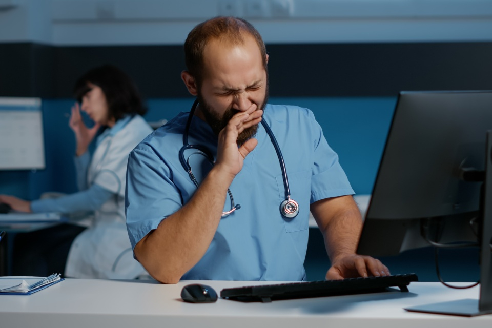 Un homme en blouse médicale bleue assis devant un ordinateur, bâillant