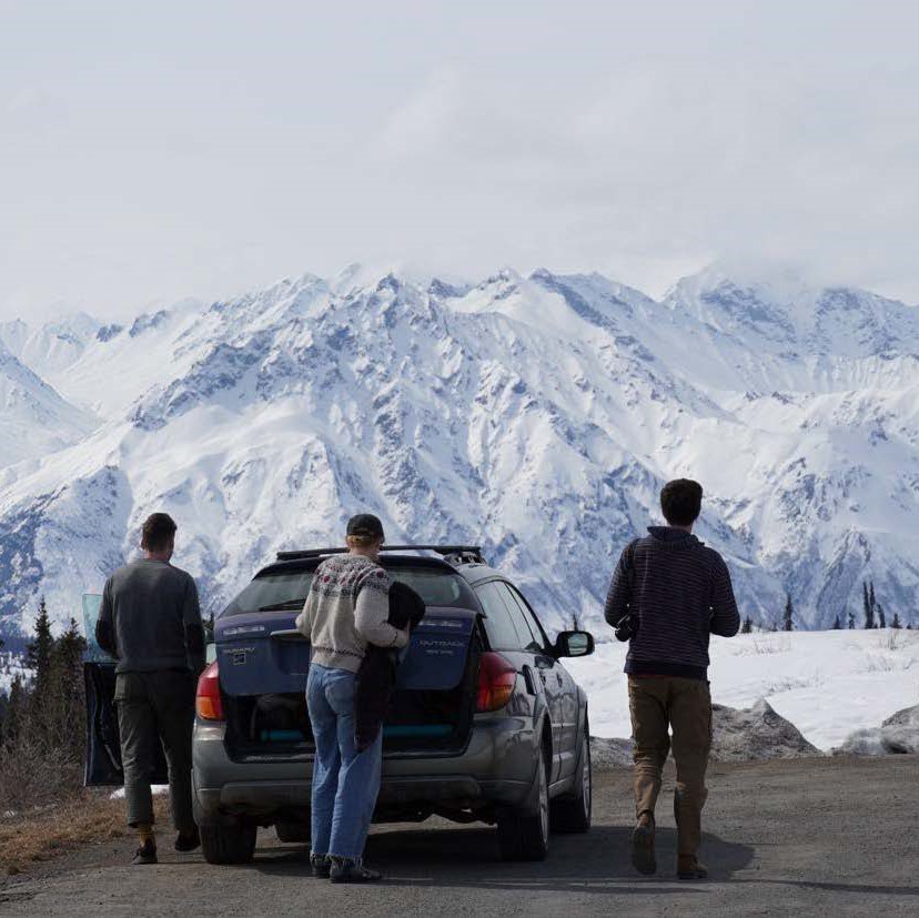 Three people stand in front of their car, looking out at a beautiful snowy mountainscape.