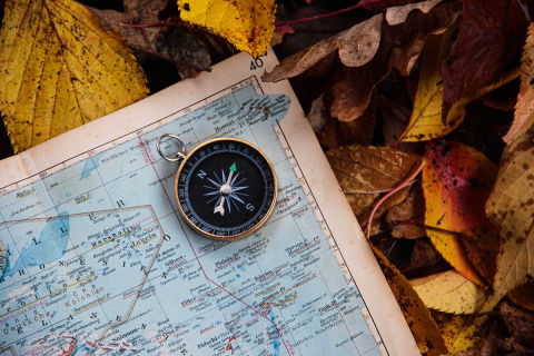 An antique compass rests on a map lying on a bed of dry leaves