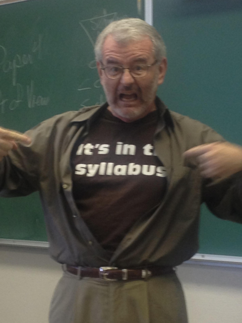 Teacher with exasperated expression points to "It's in the syllabus" message on his shirt