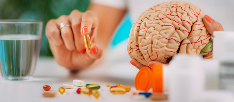 A person holding a supplement capsule next to a model of a human brain, with various pills and a glass of water on the table.