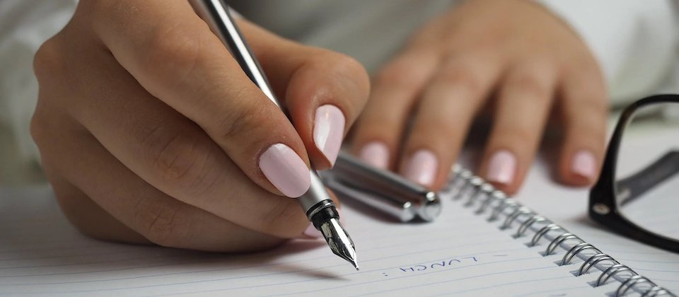 Woman in White Long Sleeved Shirt Holding a Pen Writing on a Paper