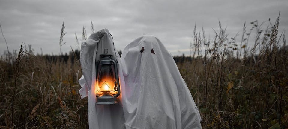 Person in ghost costume holding up lantern in a field