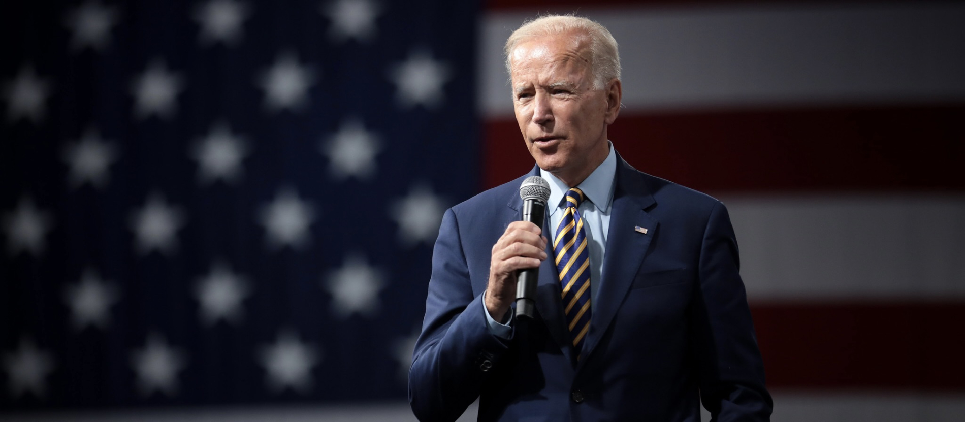 Former President Joe Biden speaking into a microphone in front of a large American flag backdrop