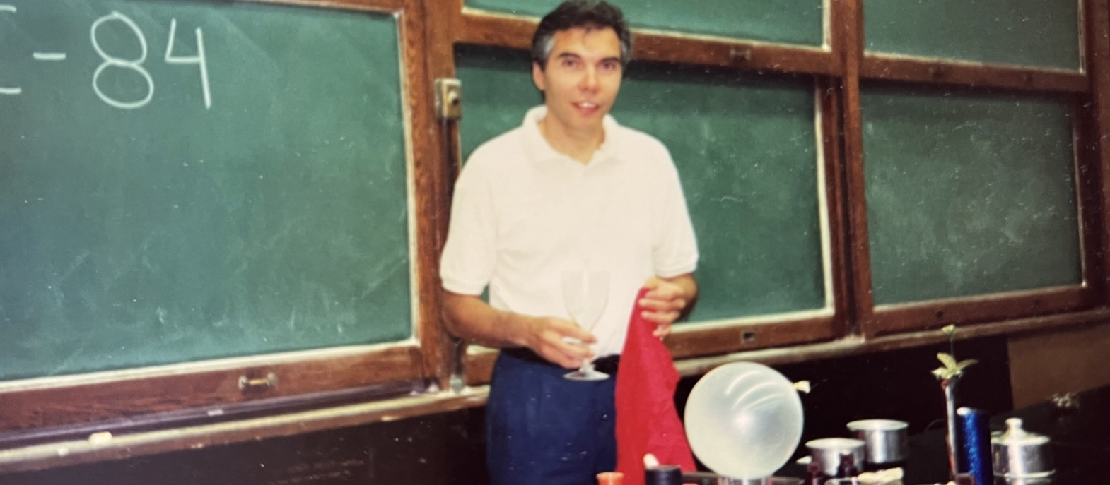Photo of Dr. Joe Schwarcz in a classroom, standing by a chalkboard with a chemistry experiment on the table