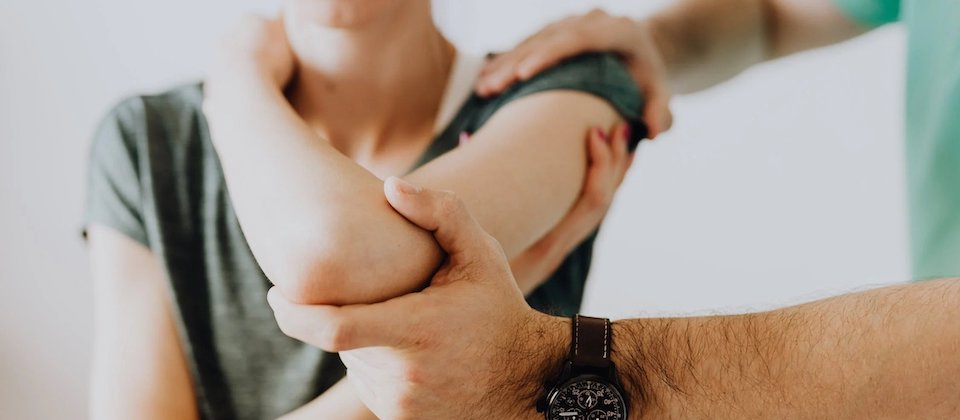 Patient with left arm bent with hand on opposite shoulder. Second person holding the patient's arm at the shoulder and elbow. Appears to be guiding through movement or stretch.