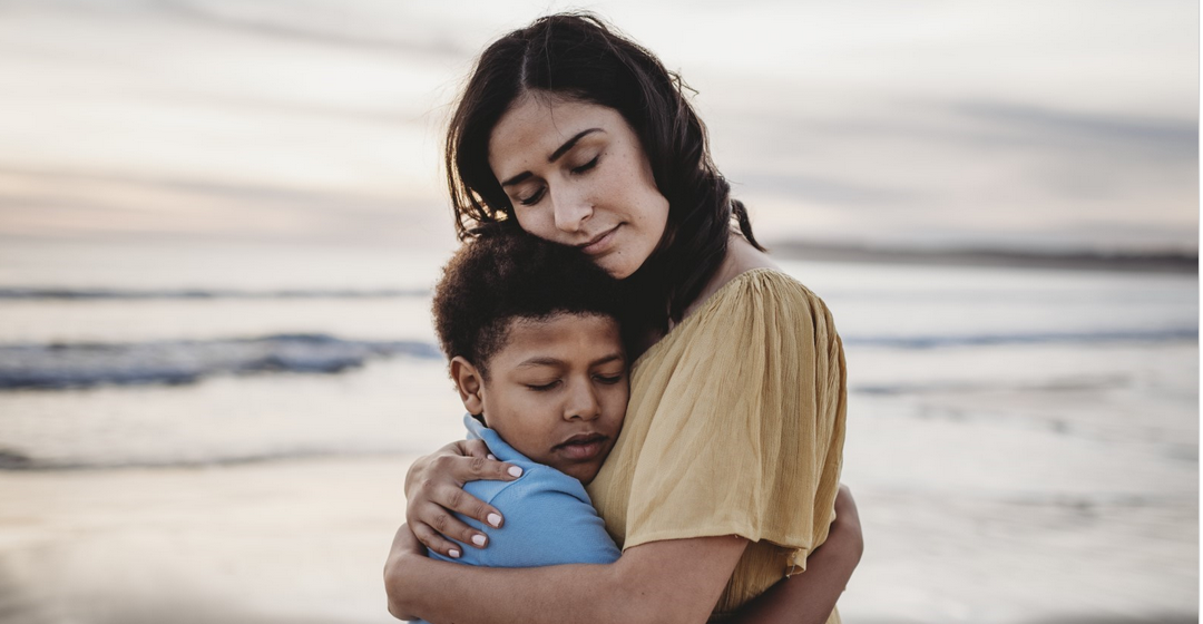 A woman hugs a child. There is a gray ocean in the background.