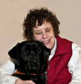 Une femme aux cheveux noirs et bouclés sourit et serre dans ses bras un gros chien noir.