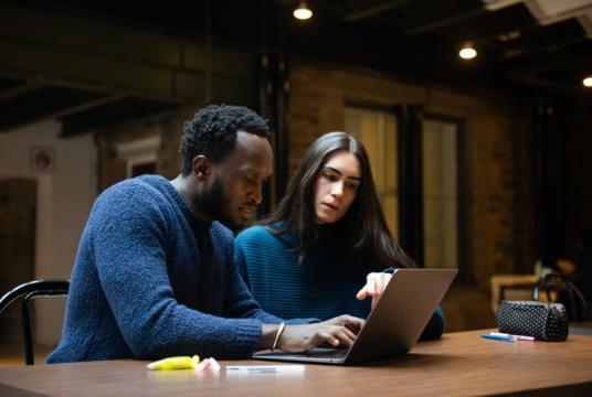 Two people sitting together at a table looking at a laptop screen