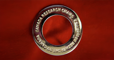 A silver circle broach with the words "Canada Research Chair" inscribed. Broach is pictured on a red background. / Broche en forme de cercle en argent portant les mots "Chaire de recherche du Canada". La broche est représentée sur un fond rouge. 
