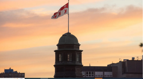 The cupola of the McGill Arts Building with an orange sky sunset behind it. The McGill flag is flying atop the cupola.  