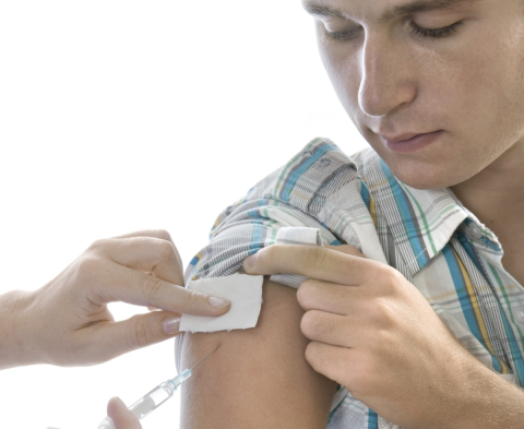 A man receives an injection in his upper arm. He is wearing a short sleeved shit. He is looking down, at the injection site. 