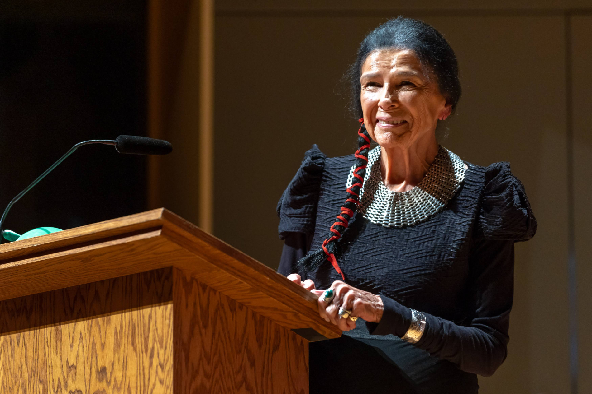 A woman (Alanis Obomsawin) stands at a podium, smiling. She is wearing a black dress. Her hair is braided and she is wearing a large silver necklace. 