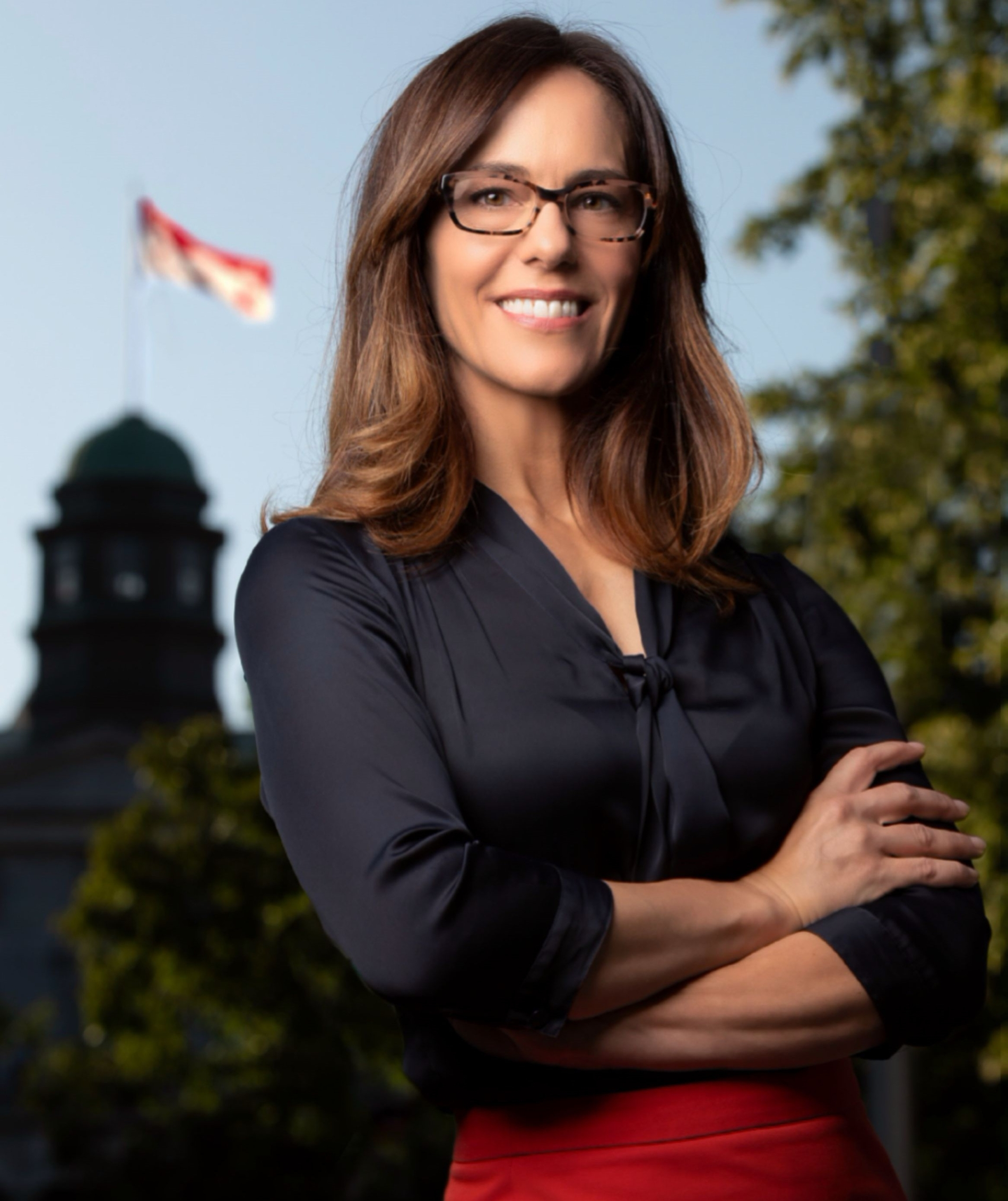 A woman stands with crossed arms in front of the McGill University campus Arts Building, She is wearing glasses and is smiling. The sky is blue. 