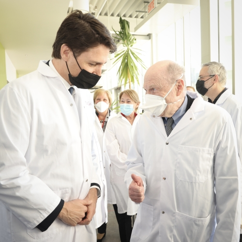 Two men - Prime Minister Trudeau and Nahum Sonenberg - talk in a hallway while wearing lab coats. 