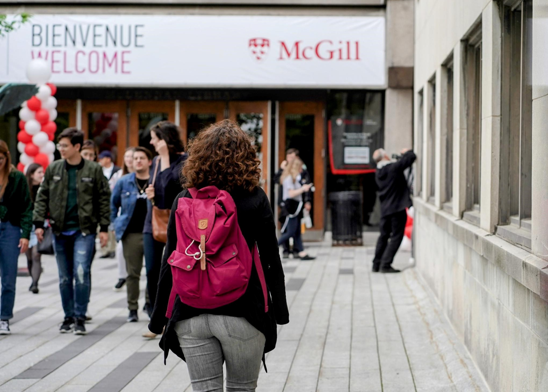 Woman wearing a red backpack walking towards a building with a banner with the McGill logo and the words 'Welcome' 'Bienvenue' 