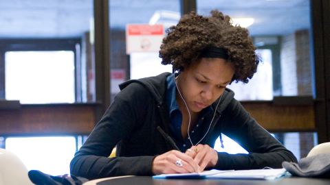 Student in black hoodie writing in notebook while listening to music