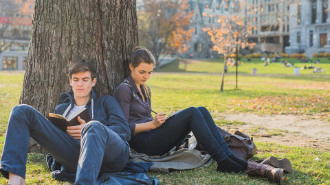 Two students sitting outside on the ground against a tree on campus